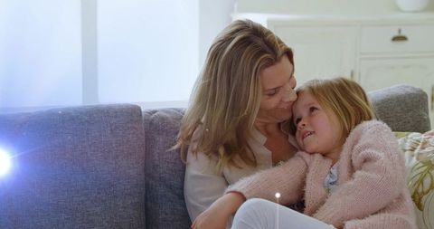 Mother cradling daughter on sofa sharing tender cuddle in pastel-lit cozy living room