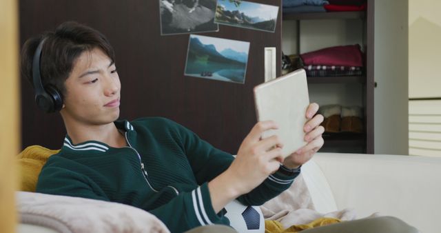 Young Asian man is relaxing on a sofa in a cozy living room, wearing headphones and holding a tablet. Illustrates comfort, leisure time, and engagement with technology. Ideal for use in ads or promotions related to home lifestyles, digital devices, or entertainment services.