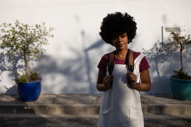 This image features a happy biracial woman standing on a sunny city street, wearing a white dress and a backpack. She is looking at the camera with a smile. The background includes potted plants and a white wall, creating a casual and urban atmosphere. This image can be used for lifestyle blogs, travel websites, fashion promotions, or advertisements focusing on urban living and casual fashion.