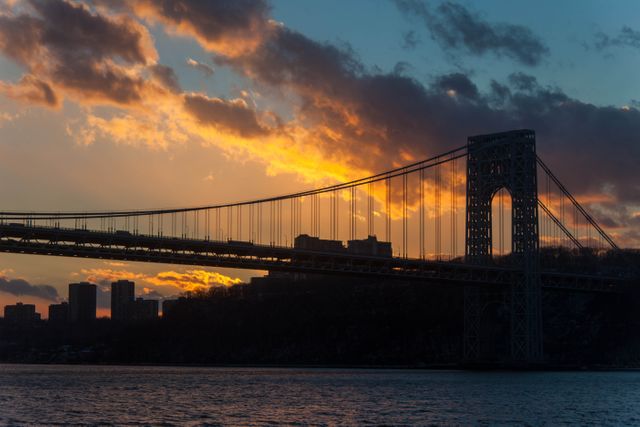 Sunset casting a golden glow on a towering suspension bridge with a city skyline background, framed by water and a dramatic sky with clouds. Perfect for urban tourism promotions, landscape photography prints, and articles about architectural landmarks or sunset travel destinations.