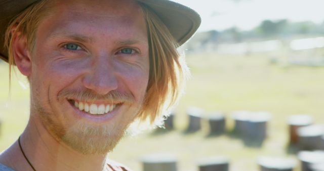 Image shows a close-up of a young man with blond hair and a hat, smiling brightly outdoors on a sunny day. This image can be used in campaigns promoting outdoor activities, happiness, casual fashion, or personal stories. Ideal for marketing materials, blog posts, and website content focused on a laid-back, joyful lifestyle.