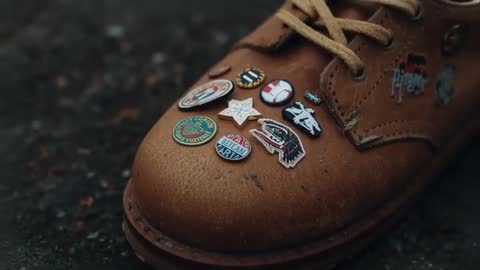 Close-up showing brown leather work boot adorned with enamel pins on wet pavement
