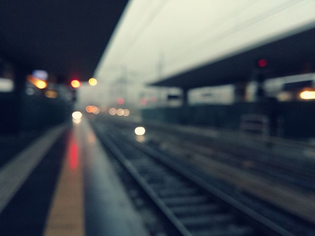 This image captures an urban train station platform on a rainy and misty day, with blurred tracks and distant lights. The blurred effect adds a sense of motion and atmosphere. Ideal for use in travel-related content, transportation articles, urban lifestyle themes, and background images for websites focusing on city life and mobility.