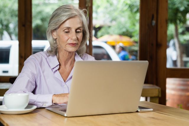 Senior woman working on laptop in a cafe, enjoying coffee. Ideal for illustrating remote work, senior lifestyle, technology use among elderly, and casual work environments.