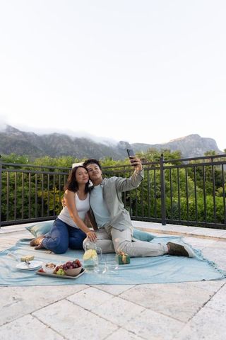 Couple taking selfie on rooftop picnic with mountain view, snacks and gift