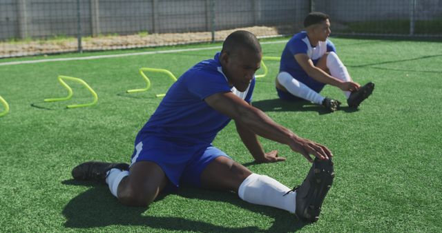 Diverse male football players wearing blue uniforms training and stretching on outdoor pitch. Football, sports, fitness and teamwork.