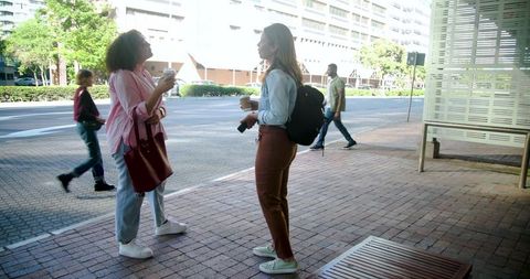 Diverse women colleagues chatting on urban sidewalk holding takeaway coffee and phones