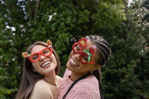 Diverse female friends laughing together in garden wearing festive novelty glasses and knit sweaters