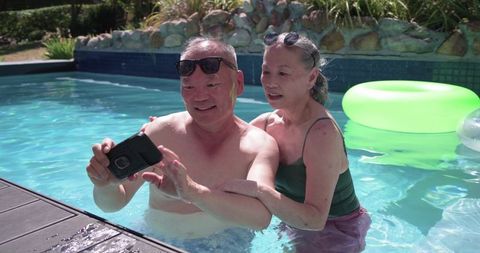 Senior couple taking pool selfie while hugging and smiling on sunny backyard float