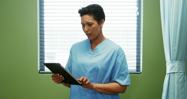 Female nurse wearing scrubs is using a digital tablet in a well-lit hospital room with green walls and a window. Useful for healthcare, medical technology, and professional healthcare staff advertisements. Ideal for illustrating modern healthcare practices and nurse-related articles.