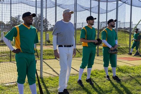 Senior coach leading mixed-age baseball players during sunny outdoor batting cage practice