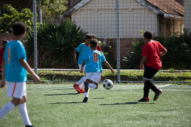 Boys from multi-ethnic soccer teams are actively playing on a green football pitch under the sun. They are passing, kicking, and shooting the ball, showcasing teamwork and competition. This image is ideal for promoting youth sports, healthy lifestyles, teamwork, and outdoor activities.