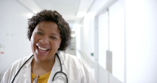 A smiling nurse in a white lab coat stands in a bright hospital hallway with a stethoscope around her neck. This image is ideal for healthcare advertisements, articles about nursing, hospital websites, and brochures promoting medical services.