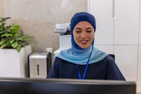 Friendly muslim receptionist wearing blue hijab and id badge at modern office front desk