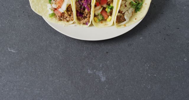 Tacos filled with ground beef, diced tomatoes, shredded lettuce, cheese, red cabbage, and onions on white plate. Gray stone background enhances the colorful and fresh ingredients. Perfect for use in food blogs, recipe books, social media promotions, or restaurant menus.