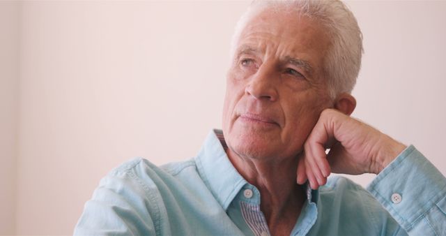 Elderly man wearing a light blue shirt sitting and looking thoughtfully into the distance. This image can be used for campaigns related to senior wellness, aging, wisdom, lifestyle of older adults, and promotional material for retirement homes, healthcare services for seniors, and articles about elderly life reflections.