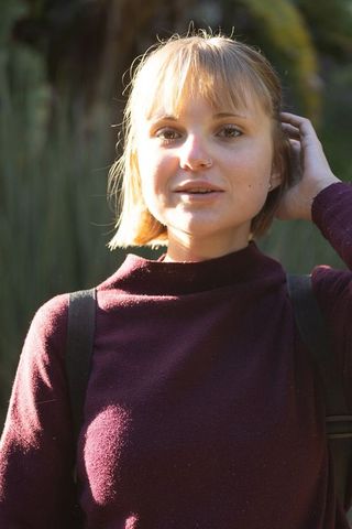 Young woman standing in sunlit garden touching hair wearing burgundy sweater, portrait