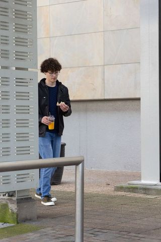 Teen walking holding orange juice and eating pastry beside modern tiled building