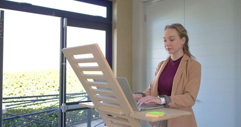 Professional woman working at wooden standing desk with laptop near sunlit glass door