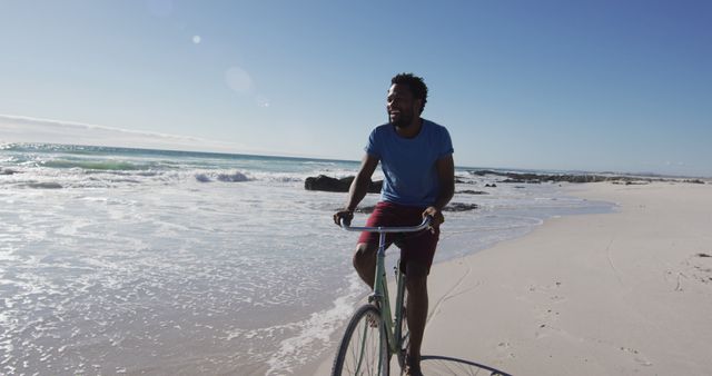 African american man smiling and riding bike on the beach. healthy outdoor leisure time by the sea.