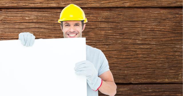 Worker wearing a yellow hardhat and gloves is holding a blank billboard, smiling cheerfully with a wood texture as a background. Ideal for construction advertisements, promotions, or safety campaigns. This image can also be used for DIY project promotions, rental business ads, or building material marketing.