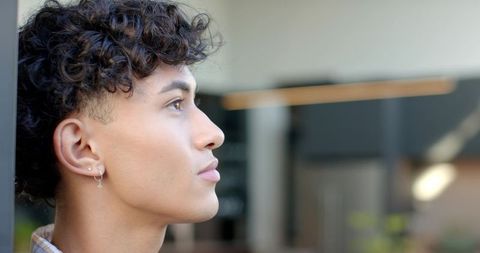 Curly-haired young man gazing in modern atrium wearing cross hoop earring, contemplative