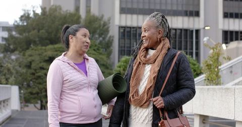 Mature Asian woman and senior African American woman walking with yoga mat in urban plaza
