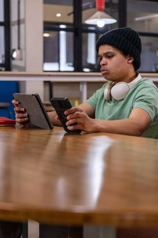 Teenage male studying on tablet and phone wearing headphones and beanie in coworking space