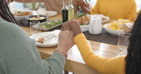 Diverse family holding hands at sunlit dining table sharing roast, corn and moments of togetherness