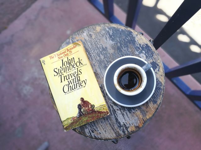 Book alongside an espresso cup on a weathered table, suggesting a peaceful reading moment. Ideal for posts about relaxation, literature, vintage aesthetics, coffee breaks, and outdoor leisure activities. Can be used in blogs about traveling, staying cozy, or enjoying simple pleasures.