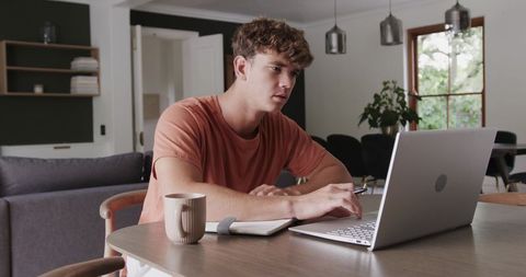Young man focusing on laptop at dining table in minimalist open-plan home office