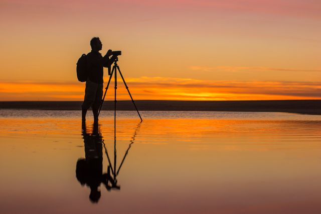 Photographer taking pictures at sunset, standing with camera on tripod by beach waters. Beautiful orange and red sky with ocean reflections. Great for travel advertisements, nature documentaries, photography magazines, serene landscape promotions, and hobbyist websites.