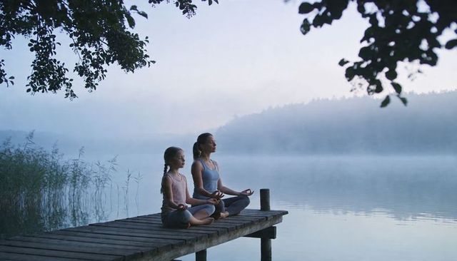 Woman and girl meditating on lakeside dock at dawn mist for mindfulness and wellness