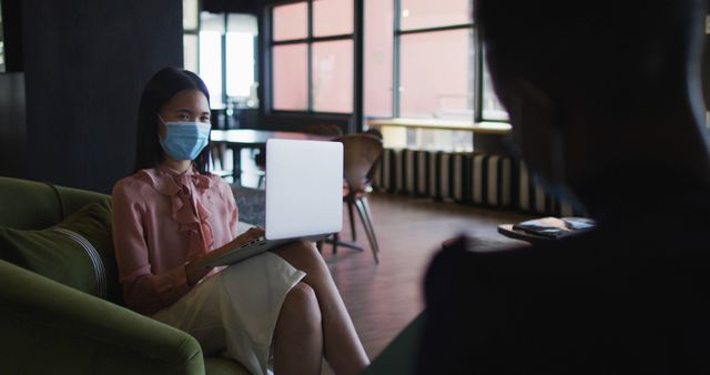 Businesswoman sitting on a couch in an office lounge, wearing a mask, working on a laptop. The image conveys themes of professional work, hygiene, and safety during a pandemic. Ideal for use in articles, blogs, or presentations discussing workplace safety, remote work, or pandemic measures in professional environments.