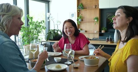Diverse women laughing and sharing meal with wine around sunlit wooden dining table