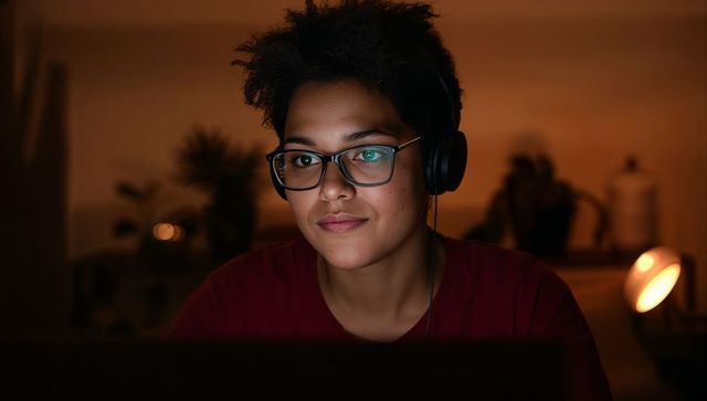 Young remote worker concentrating on laptop at night wearing headphones and glasses