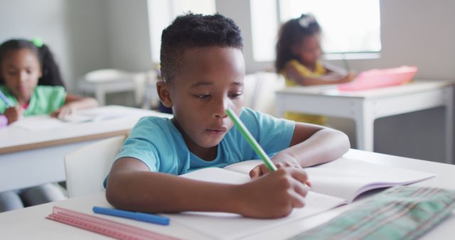 Boy writing in classroom, focused on task, with two classmates in background at their desks. Perfect for educational materials, school websites, learning resources, and academic posters to portray focus, learning, and educational settings.