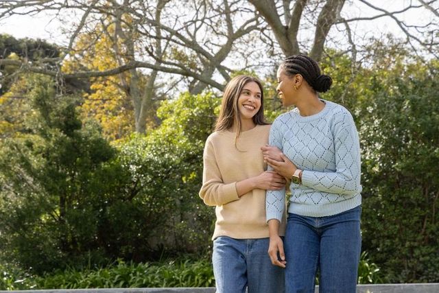 Diverse women smiling and walking arm in arm on park ledge wearing sweaters and jeans