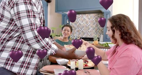 Friends sharing dinner and passing bowl in cozy kitchen with floating purple hearts