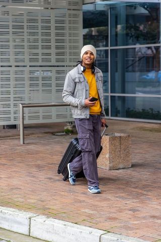 Young man walking with rolling suitcase and smartphone in urban plaza
