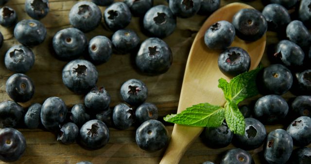 Fresh blueberries scattered around wood spoon and mint leaves on rustic table. Ideal for promoting healthy eating, organic food products, nutrition blogs, and summer recipes. Perfect for illustrating concepts of freshness, vegan diets, and natural foods.