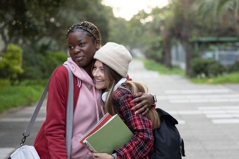 Teen friends hugging on tree-lined sidewalk holding notebooks and backpacks smiling