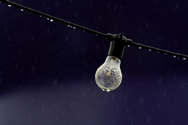 Vintage light bulb hanging on wire outdoors in rainy night with droplets creating dramatic visual effect. Useful for themes related to nostalgia, outdoor lighting design ideas, mood lighting, weather ambiance, and night photography.