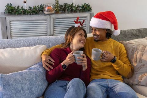 Cozy couple cuddling on sofa sharing mugs wearing santa hat and reindeer antlers