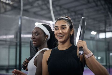 Smiling diverse female teammates holding padel paddles on indoor court celebrating teamwork