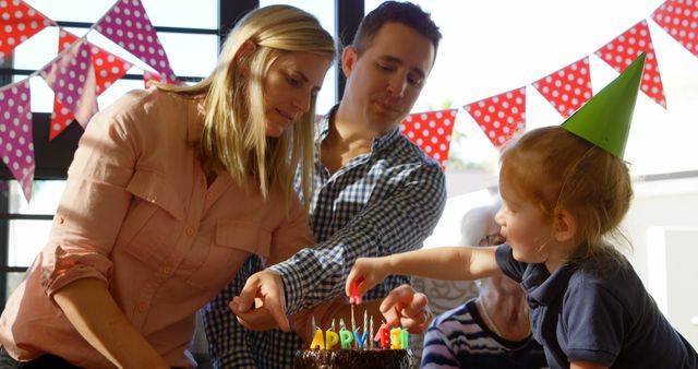 This image depicts a family celebrating a little girl's birthday. Parents assist the child in blowing out birthday candles on a chocolate cake. Vibrant birthday decorations, including colorful bunting and a party hat, set a joyful atmosphere. This can be used in articles about family celebrations, birthday party planning, and parent-child bonding.