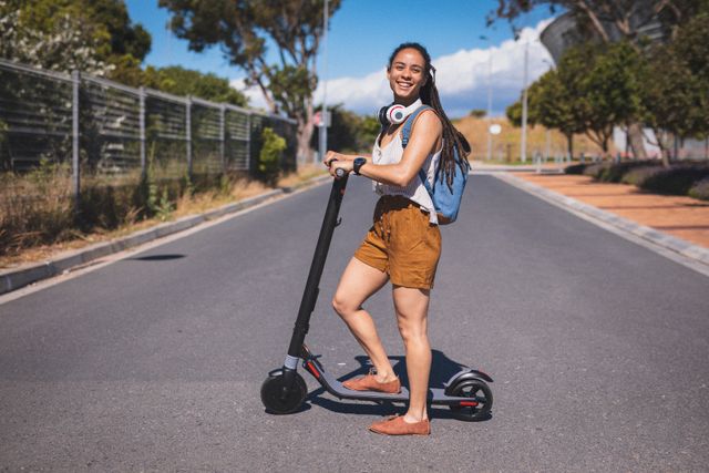 Portrait of biracial woman wearing headphones and backpack standing on scooter in street smiling. digital nomad on the go in summer.