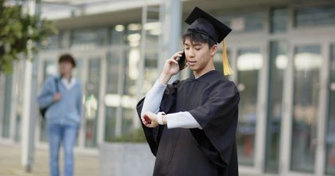Asian graduate checking smartwatch while talking on phone wearing mortarboard on campus