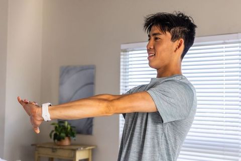 Asian man stretching arms near window blinds wearing white fitness band during home workout