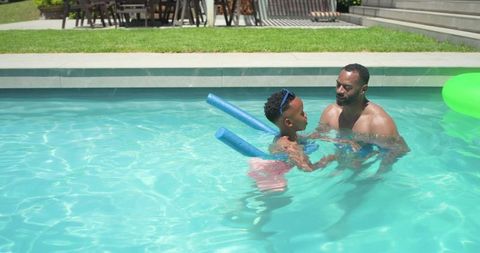 African american father teaching son to swim in backyard pool with foam noodles